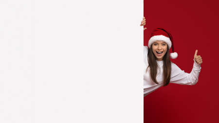 Holiday Ad. Excited Woman Wearing Santa Hat Peeking Behind White Advertisement Boardの写真素材