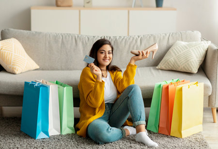Overjoyed middle eastern woman holding credit card and new shoe, sitting on floor among colorful shopper bagsの写真素材