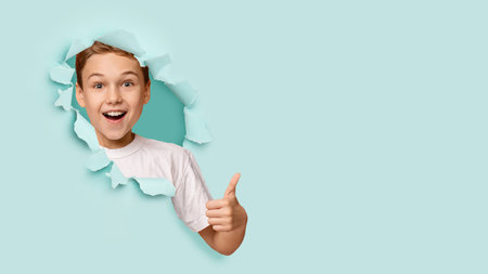 Satisfied excited caucasian teen boy with open mouth, looking through hole on green paper and showing thumb upの写真素材