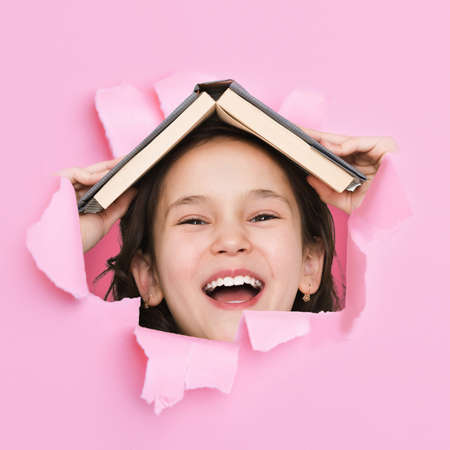 Happy excited pretty cute asian little girl with open mouth and books on her head looks through hole on pink paperの写真素材