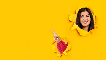 Smiling millennial spanish woman looks through hole in yellow paper, points her finger at empty space, collageの写真素材