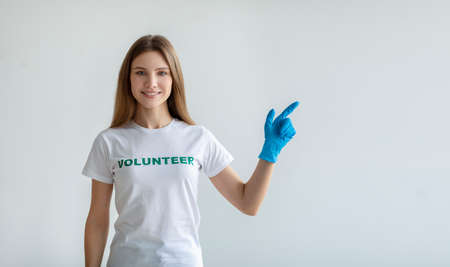 Look here. Happy female volunteer in gloves pointing aside at free space, standing over light wall and smiling at cameraの写真素材