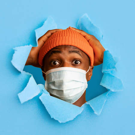 Surprised millennial african american guy with wide open eyes in protective mask holds his head with handsの写真素材