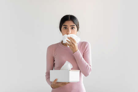 Shocked upset young hindu woman suffering from flu, blows nose, cough, holding box of napkinの写真素材