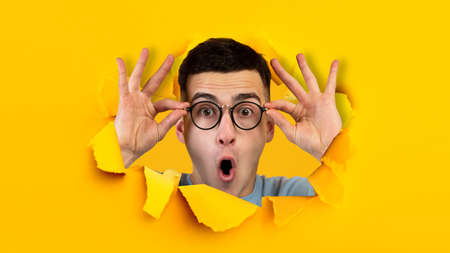 Shocked positive young european guy with open mouth, touches glasses, looks through hole in yellow paperの写真素材