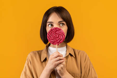 Sweet tooth concept. Playful young caucasian lady holding lollipop, closing and covering mouth with...