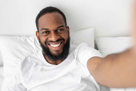 Smiling millennial african american bearded man looks at webcam, lies on white bed in bedroom...