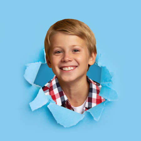 Smiling european handsome blond teen boy pupil looking through hole on blue paper, free space, squareの写真素材