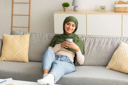 Peaceful weekend. Positive young Arab lady in hijab relaxing on sofa with cozy hot drink at home