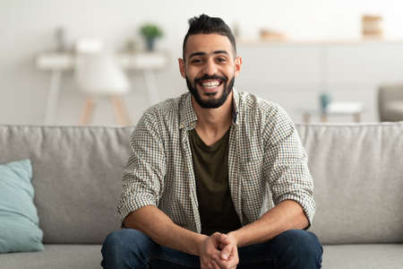 Portrait of handsome young Arab man smiling and looking at camera, sitting on sofa at homeの写真素材