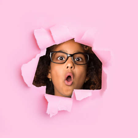 Shocked funny african american little girl in glasses with open mouth looks through hole on pink paperの写真素材