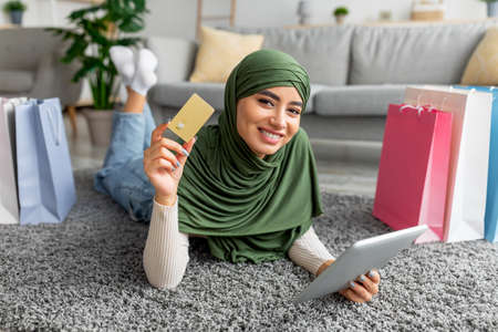 Happy Arab lady in hijab lying on floor with digital tablet, paper bags and credit card, shopping on...