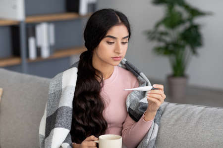 Upset young hindu woman with cup of hot drink in plaid, suffering from cold, sits on sofa in living roomの写真素材