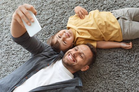 Happy family portrait. Young father doing selfie with his little son, smiling to smartphone, resting together on floorの写真素材