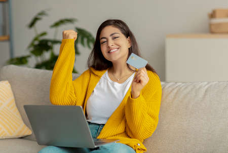 Overjoyed young arab lady with credit card and laptop winning in online lottery, shopping on huge...