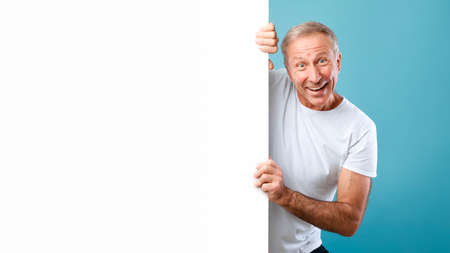 Mature man peeking out blank white advertising billboard at studioの写真素材