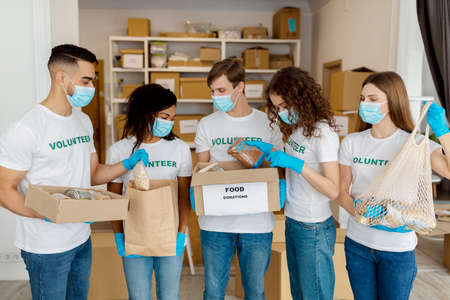 Charity center. Group of diverse young people in medical masks packing donation boxes, preparing...