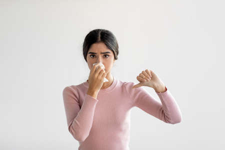 Upset unhappy young indian lady sneezes in napkin, blows her nose and shows thumb down gesture