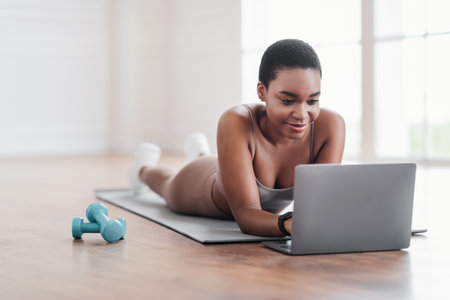 Smiling sporty black woman lying on yoga mat using laptopの写真素材