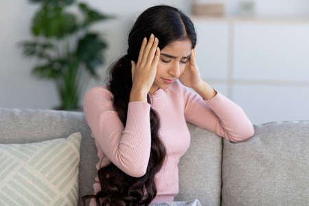 Despaired upset young hindu female suffer from headache and flu, sits on sofa and holds her head with handsの写真素材