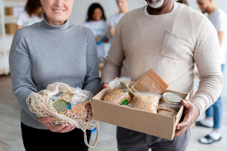 Mixed race elderly spouses showing donation food in box and string bag, volunteers packing boxes for poor peopleの写真素材