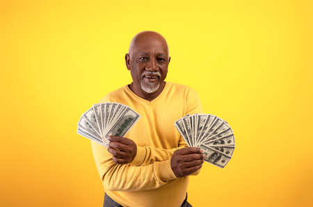 Happy rich senior black man holding two fans of money, celebrating big financial luck and success on orange backgroundの写真素材