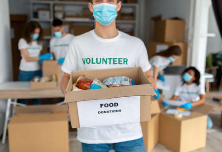 Charity organization. Male volunteer in medical mask, holding box with food donations, closeup, croppedの写真素材