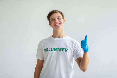 Volunteer work. Happy activist guy in white volunteer t-shirt showing thumb up, posing and smiling over light backgroundの写真素材