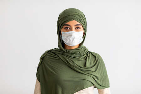 Pretty millennial Arab female in hijab wearing medical mask during coronavirus epidemic on light studio backgroundの写真素材