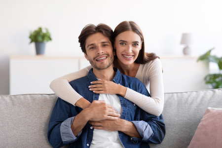 Happy young wife hugs husband from behind on sofa and look at camera in room interiorの写真素材