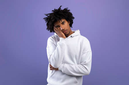 Portrait of black teen guy feeling bored, leaning on his hand, having dull day on violet studio...
