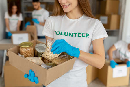 Female activist volunteer working at charity center, holding box with food donations, grains, beans and canned foodの写真素材