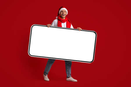 Young Cheerful Man In Santa Hat Holding Big Blank Smartphone In Hands