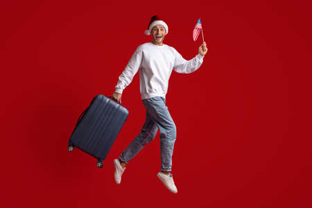 Excited Young Man In Santa Hat Jumping With American Flag And Suitcaseの写真素材