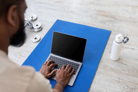 Young black guy typing, watch lesson on laptop with blank screen on mat on floor with bottle of waterの写真素材