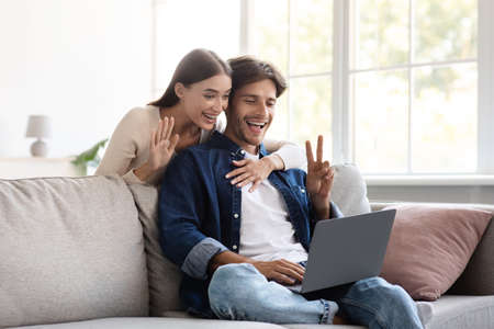 Smiling millennial european couple communicate with family remotely, wave hand, showing peace signの写真素材