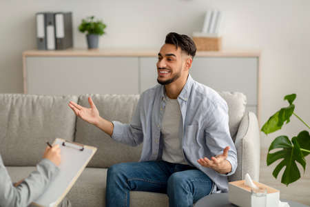 Young happy arab guy talking to his psychologist, receiving professional help, sitting on sofa at mental health clinicの写真素材