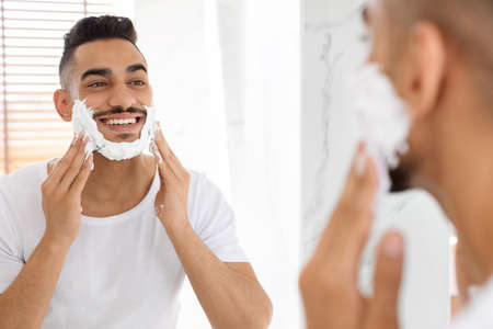 Handsome Arab Man Applying Shaving Foam On Face Near Mirror In Bathroomの写真素材