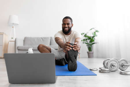 Glad young black bearded male athlete do leg stretches on mat on floor in room interior with...