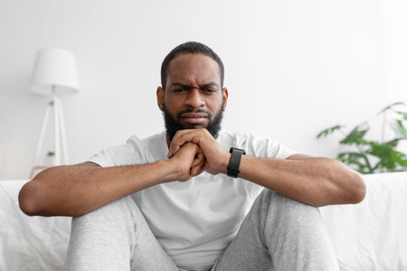Sad pensive young african american bearded male sitting in room interior, on white wall backgroundの写真素材
