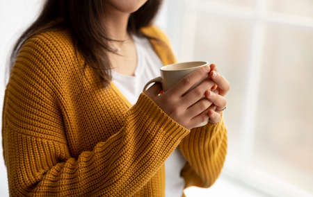 Cropped view of arab woman warming her hands on cup of hot coffee near window at home, empty spaceの写真素材