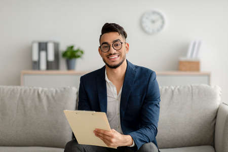 Portrait of happy arab male psychologist smiling at camera and taking notes during therapy session at clinicの写真素材