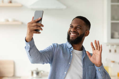 Cheerful millennial african american bearded guy waving hand to smartphone webcam, communication with followerの写真素材