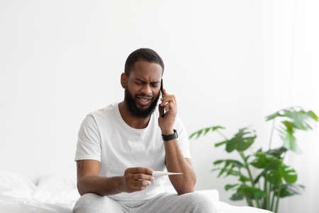 Despaired young black bearded male watch at thermometer with high fever, calling by phone