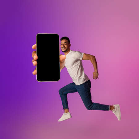 Full length portrait of happy young Arab guy jumping with smartphone, showing empty screen in neon...