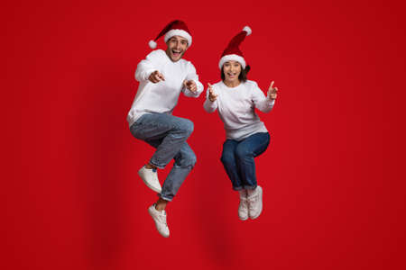 Overjoyed Man And Woman In Santa Hats Jumping And Pointing At Camera