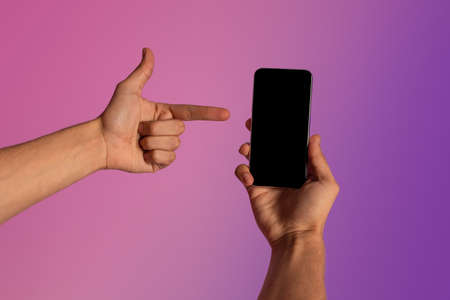 Unrecognizable young guy pointing at smartphone with empty screen in neon light. Mockup for ad, mobile app or websiteの写真素材