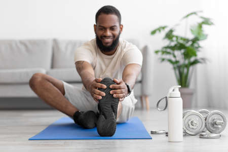 Cheerful millennial black bearded man do leg stretch on floor on mat in living room interior
