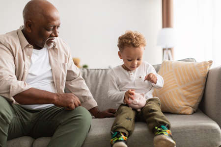 Black Grandfather And Grandchild Boy Putting Coin In Piggybank Indoor