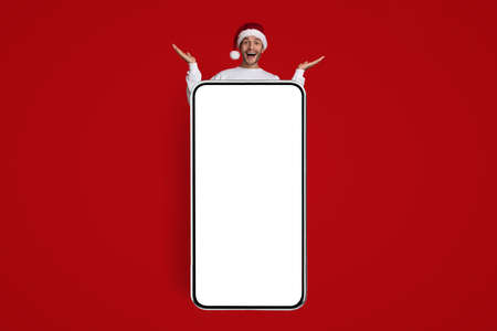 Online Offer. Funny Man In Santa Hat Standing Behind Big Blank Smartphoneの写真素材